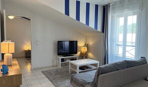 Ein Wohnzimmer mit Fernseher, Sofa und einem kleinen Tisch. Ein Fenster mit Vorhängen, eine Lampe und eine Tür zu einem anderen Raum sind ebenfalls vorhanden.