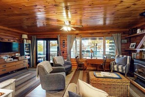 A log cabin interior with a fireplace, a television, a dining area, and a large window offering a view of the outdoors. - 2BR lakefront with hot tub, dock, & 50ft shoreline (Dundee)