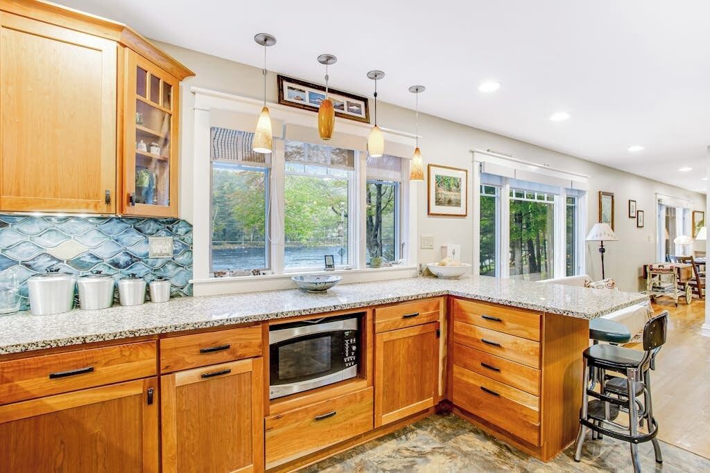A spacious kitchen with wooden cabinets, a granite countertop, and large windows offering a view of the outdoors.