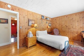 A bedroom with a wooden paneled wall, a bed with white bedding, a nightstand with a lamp, and a chair. - Beautiful 3BR Riverfront Adirondacks - Hot Tub (Chestertown)