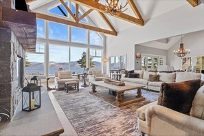 A spacious living room with a large window, a fireplace, and a high ceiling with exposed beams. - 5BR panoramic lake acreage - sauna, game tables (Bolton Landing)