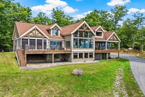 A two-story house with a stone foundation, large windows, and a deck. - 5BR panoramic lake acreage - sauna, game tables (Bolton Landing)