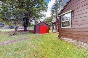 A wooden house with a red garage, a covered porch, and a large tree in the yard. - 2BR dog friendly cottage with a wood stove (North Creek)