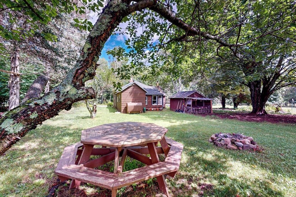 Une table de pique-nique en bois, placée dans un espace herbeux, avec une maison et un abri de jardin en arrière-plan.
