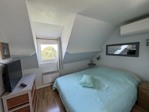 A bedroom with a sloped ceiling, a bed with a quilt, a television on a stand, a window with curtains, and a wall-mounted mirror. - Large sea view house near beach & boating (Honfleur)