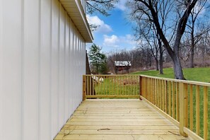 A wooden deck with a railing, a white building, and a grassy area with trees in the background. - 1BR - loft withexpansive lake views, deck & grill (Dundee)