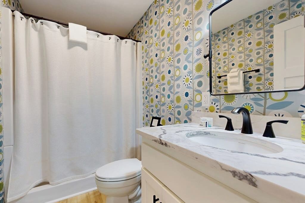 A bathroom with a shower curtain, toilet, sink with a mirror, and patterned wallpaper.