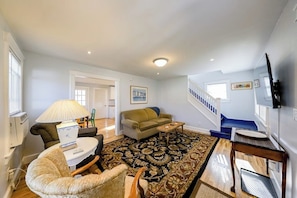 Ein Wohnzimmer mit einer Couch, einem Sessel, einem Couchtisch und einem gemusterten Teppich. Eine Treppe mit blauer Geländer führt in einen anderen Raum.