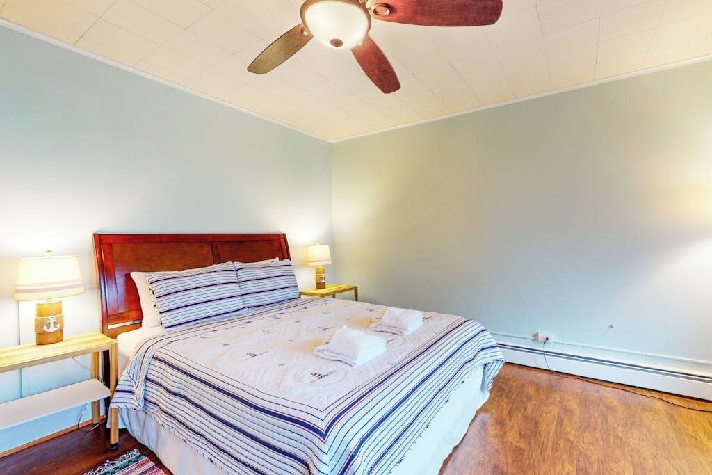 A bedroom with a wooden headboard, a bed with striped bedding, two bedside lamps, a ceiling fan, and a wooden floor.