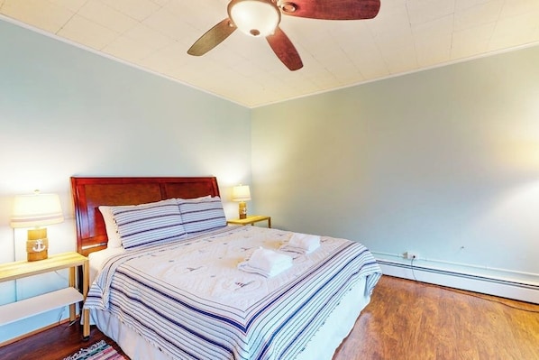 Un dormitorio con cabecera de madera, una cama con ropa de cama a rayas, dos lámparas de noche, un ventilador de techo y suelo de madera.