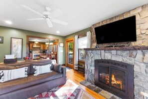 A living room with a stone fireplace, a flat-screen TV mounted above it, a ceiling fan, and a kitchen visible through an open doorway. - Scenic lakefront 4BR with water access & dock (Kattskill Bay)