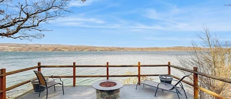 A wooden deck with a stone fire pit, wicker furniture, and a view of a body of water.