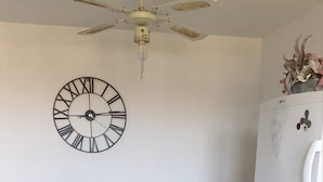Eine kompakte Küche mit Holztisch, zwei Stühlen, Deckenventilator, Wanduhr und einem weißen Kühlschrank.