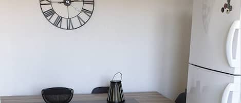 Une cuisine de taille réduite, avec une table en bois, deux chaises, un ventilateur de plafond, une horloge murale et un réfrigérateur blanc.