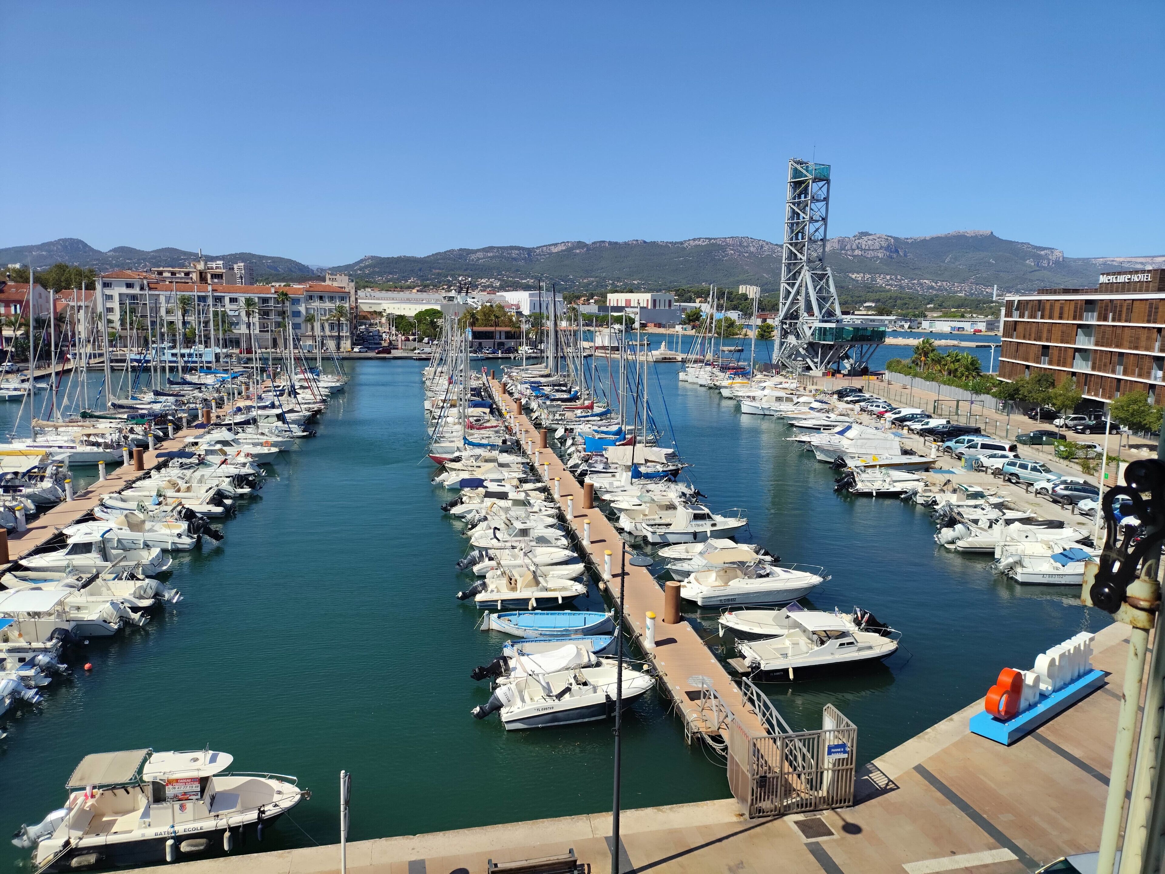 A marina with numerous boats docked, a clear sky, and a mountainous backdrop.