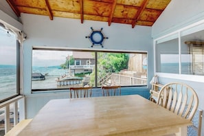 A room with a wooden table and chairs, a large window with a view of the sea and a dock, and a ship's wheel decoration on the wall. - 2BR lakefront home with grill - great views (Jerusalem)