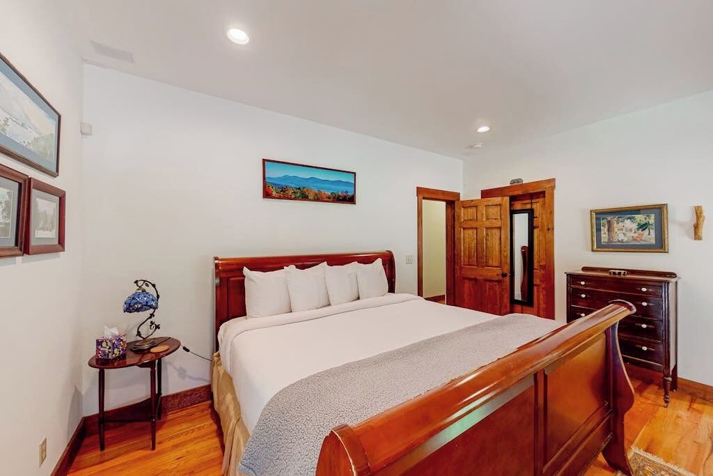 A bedroom with a wooden bed, a bedside table with a lamp, a dresser, and a door leading to another room.