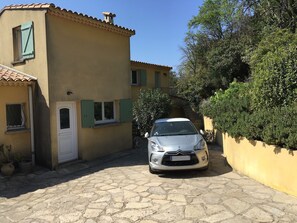 Ein silbernes Auto parkt in einer schmalen Straße, flankiert von einem gelben Gebäude und einem grünen Baum.