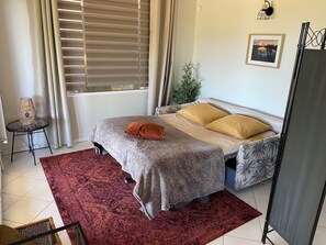 Ein Schlafzimmer mit einem Bett, einem roten Teppich, einem Sessel, einer Pflanze und einem Bild an der Wand.