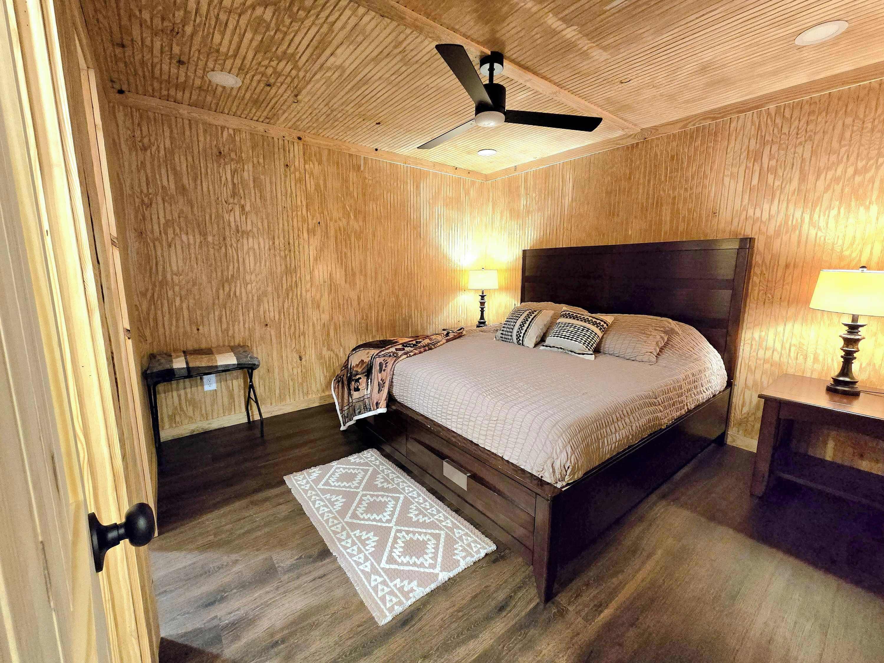 Een slaapkamer met houten lambrisering, een bed, twee bedlampen, een plafondventilator en een kamerkleed met een patroon.