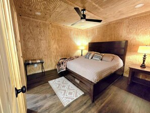 A wooden-paneled bedroom with a bed, two bedside lamps, a ceiling fan, and a patterned rug. - The Lodge at Welch-The Cabin (Welch)