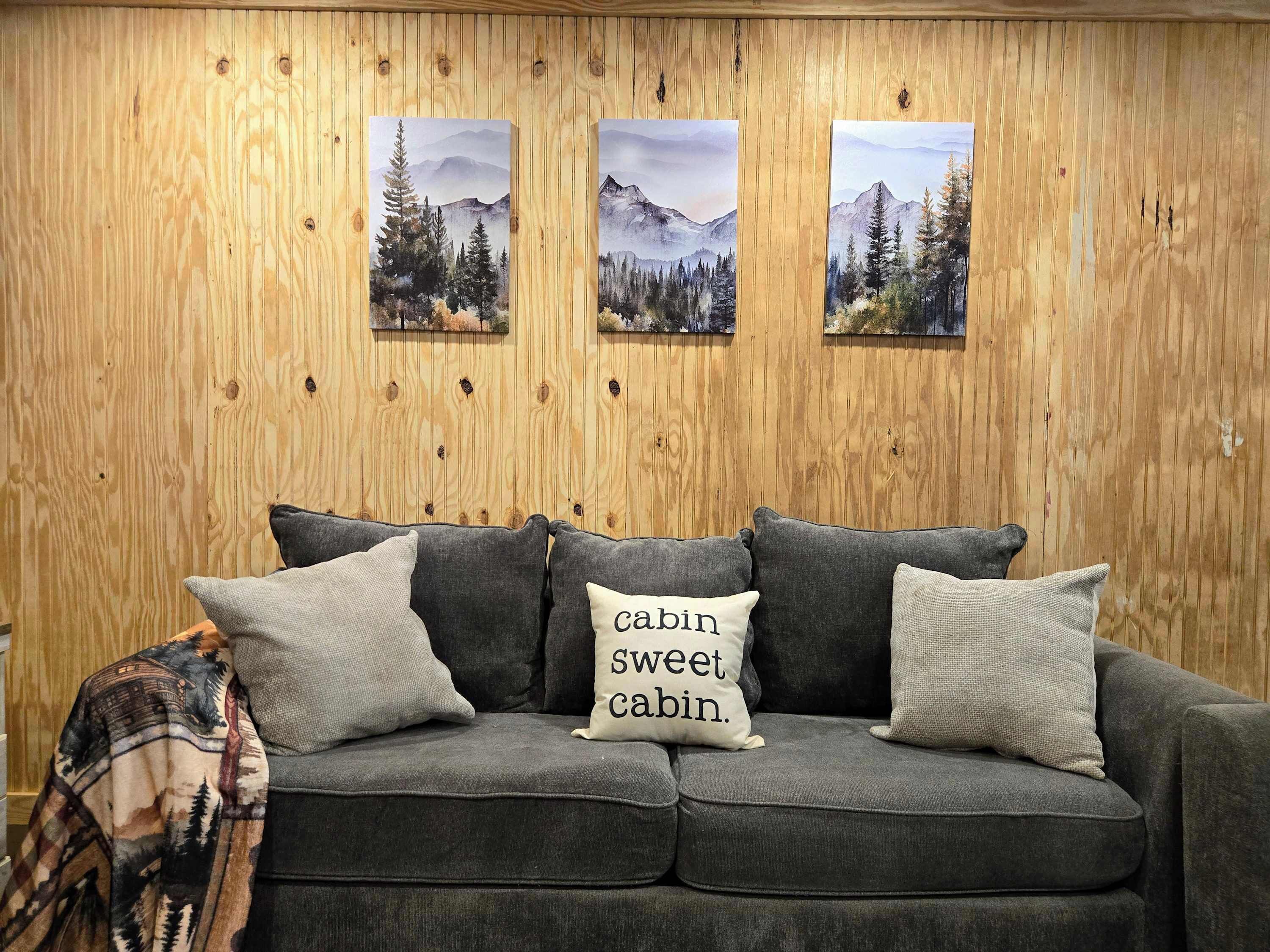 Um sofá cinzento com três almofadas, uma delas com a inscrição 'cabin sweet cabin', em frente a uma parede de madeira com três quadros de paisagens emolduradas.