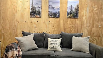 Um sofá cinzento com três almofadas, uma delas com a inscrição 'cabin sweet cabin', em frente a uma parede de madeira com três quadros de paisagens emolduradas.
