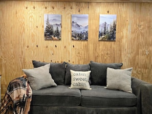 A grey sofa with three cushions, one with 'cabin sweet cabin' text, against a wooden wall with three framed landscape paintings.