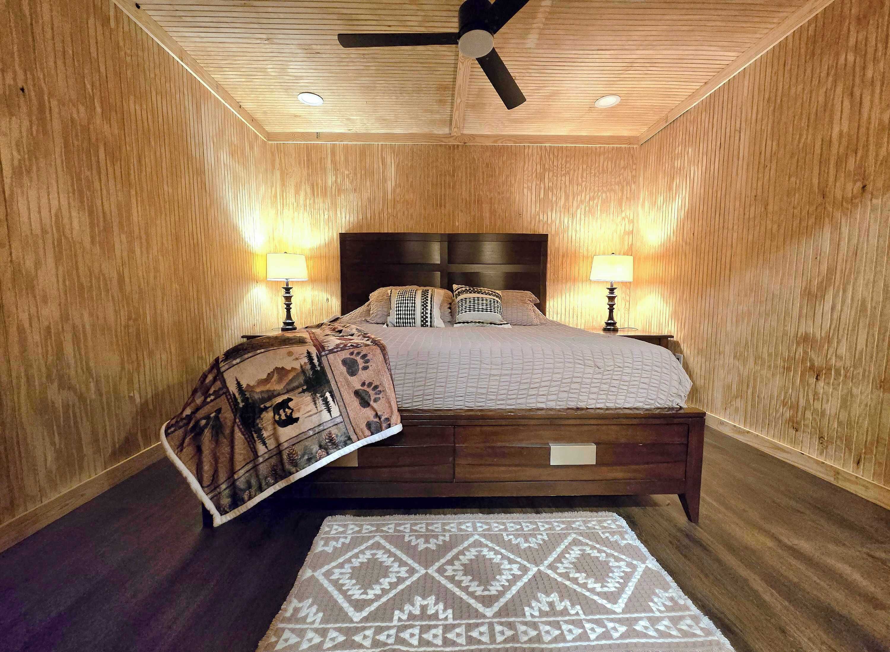 A bedroom with a wooden paneled ceiling, a bed with a headboard, two bedside lamps, and a patterned throw blanket.