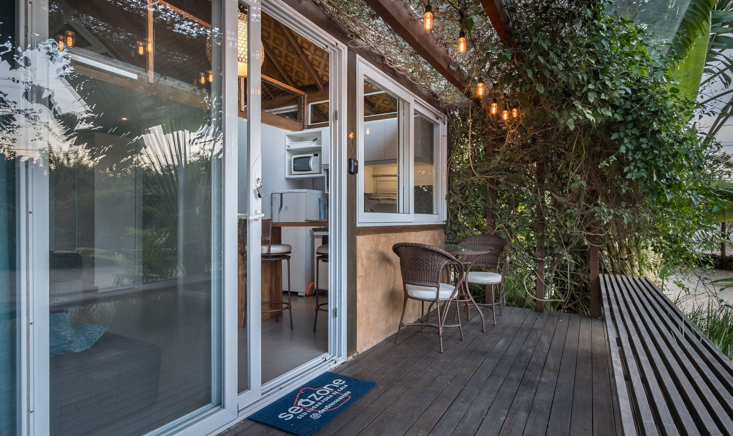 A covered outdoor seating area with wicker chairs and a table, a glass door entrance, and a doormat with text.