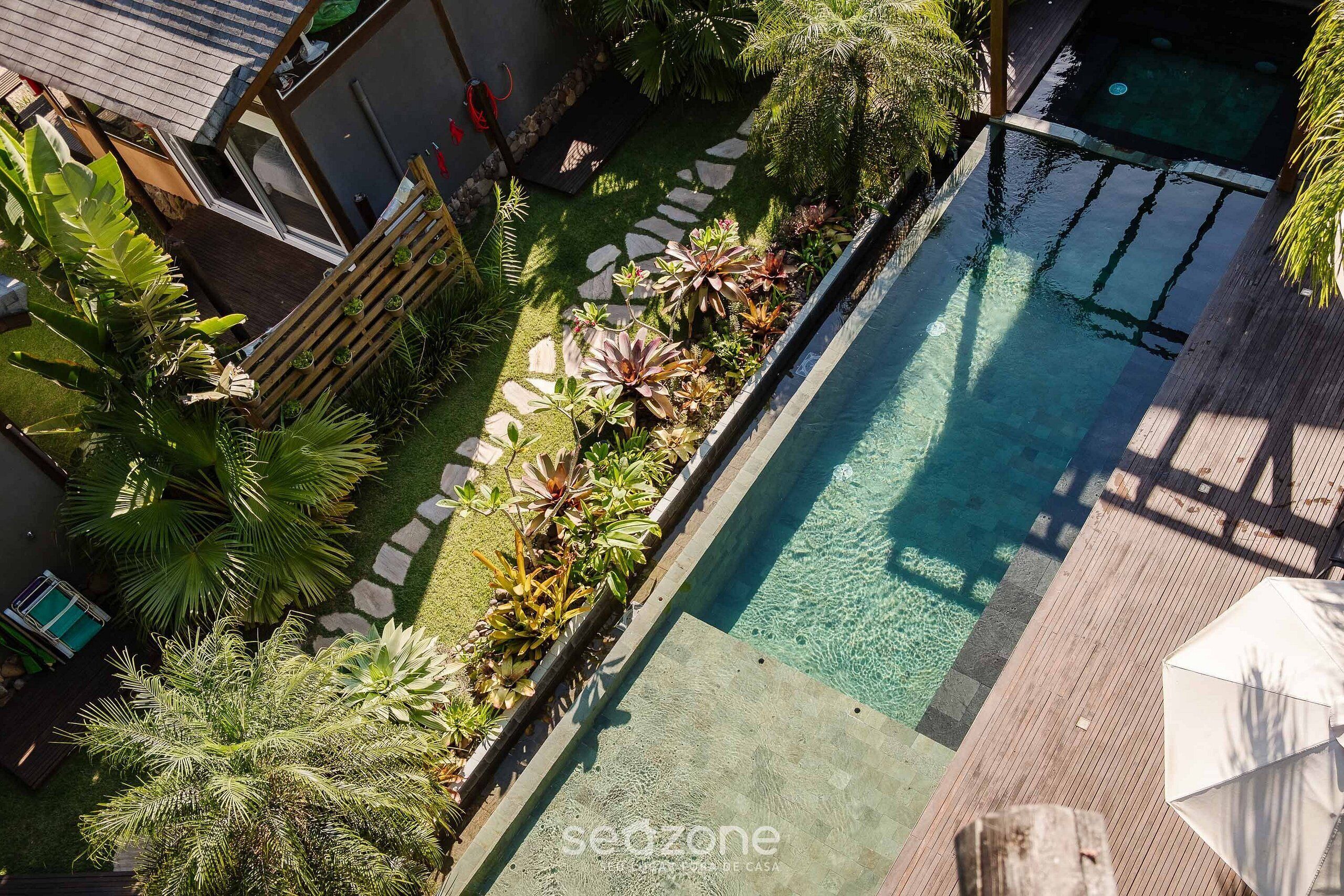 A swimming pool with a wooden deck, surrounded by lush greenery and a stone pathway.