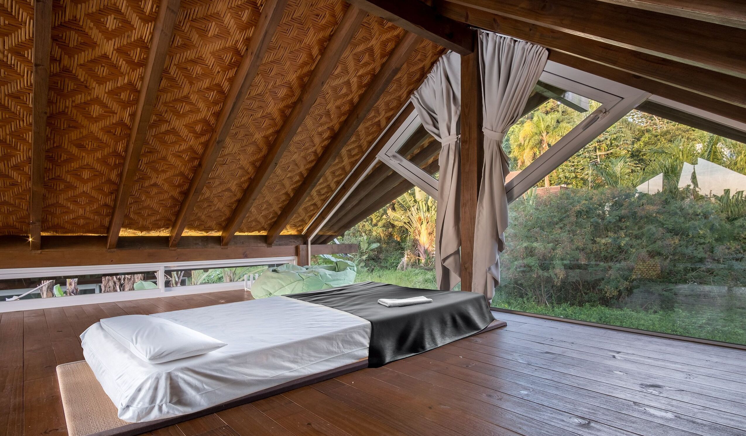 A bedroom with a wooden ceiling, a bed with a white mattress, and a view of greenery outside.
