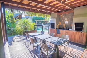 An outdoor patio with a wooden deck, tables, chairs, a barbecue area, and a stone wall. - Beautiful Cabin Barra Ibiraquera and Rosa Vgz0005 (Imbituba)