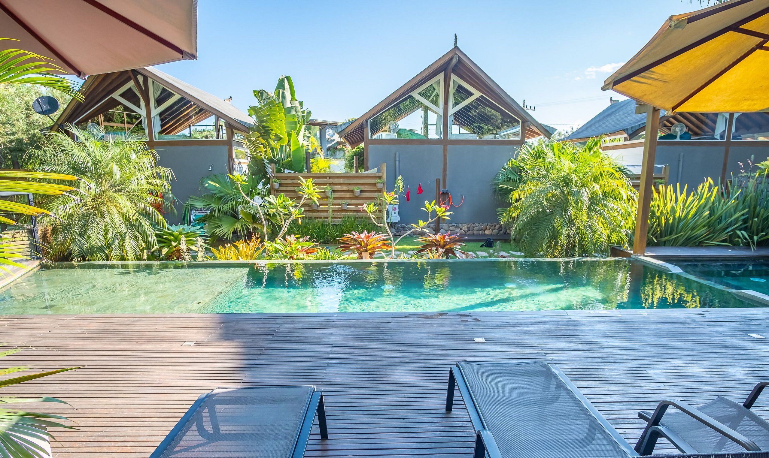 Uma área de piscina com deck de madeira, espreguiçadeiras e plantas tropicais.