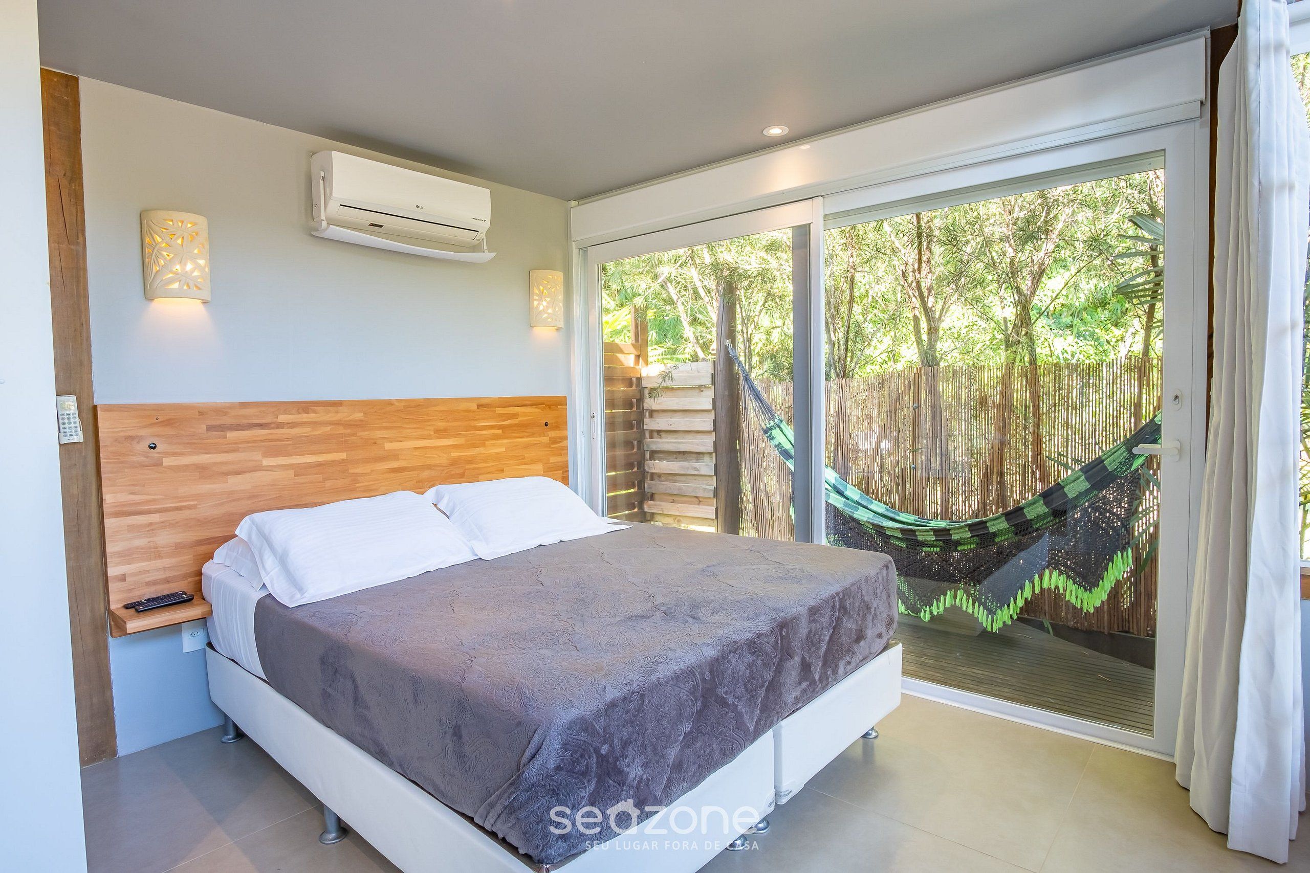 Quarto com cama, ar condicionado, mesa de cabeceira e vista para uma rede e árvores através de uma porta de correr.
