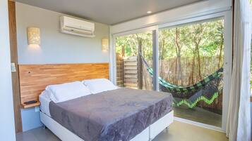 Quarto com cama, ar condicionado, mesa de cabeceira e vista para uma rede e árvores através de uma porta de correr.