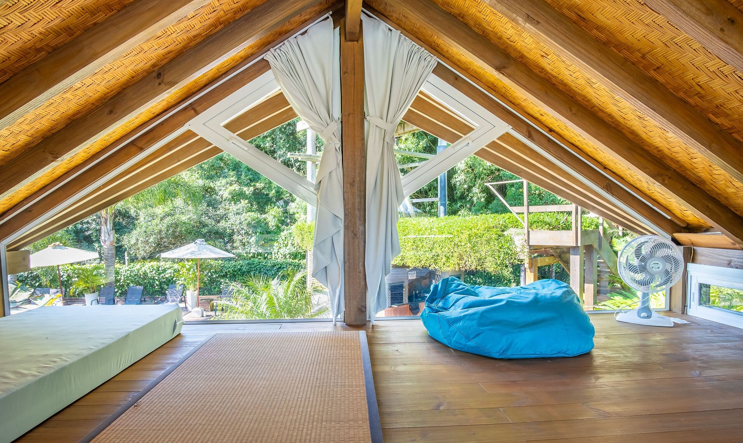 Una habitación con techo inclinado, vigas de madera, una cama, un sillón de mimbre azul y un ventilador.