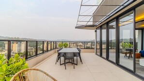 Terraço no telhado com mesa e cadeiras para refeições, cercado por paredes de vidro com vista para a cidade.