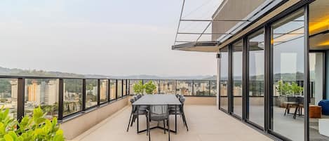Terraço no telhado com mesa e cadeiras para refeições, cercado por paredes de vidro com vista para a cidade.