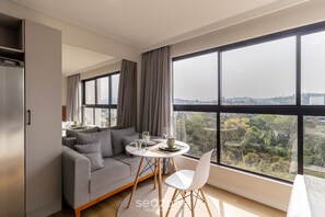 A modern living room with a grey sofa, a dining table with chairs, and a large window offering a city view. - Cozy Studio in Building With Pool Rup1405 (Blumenau)