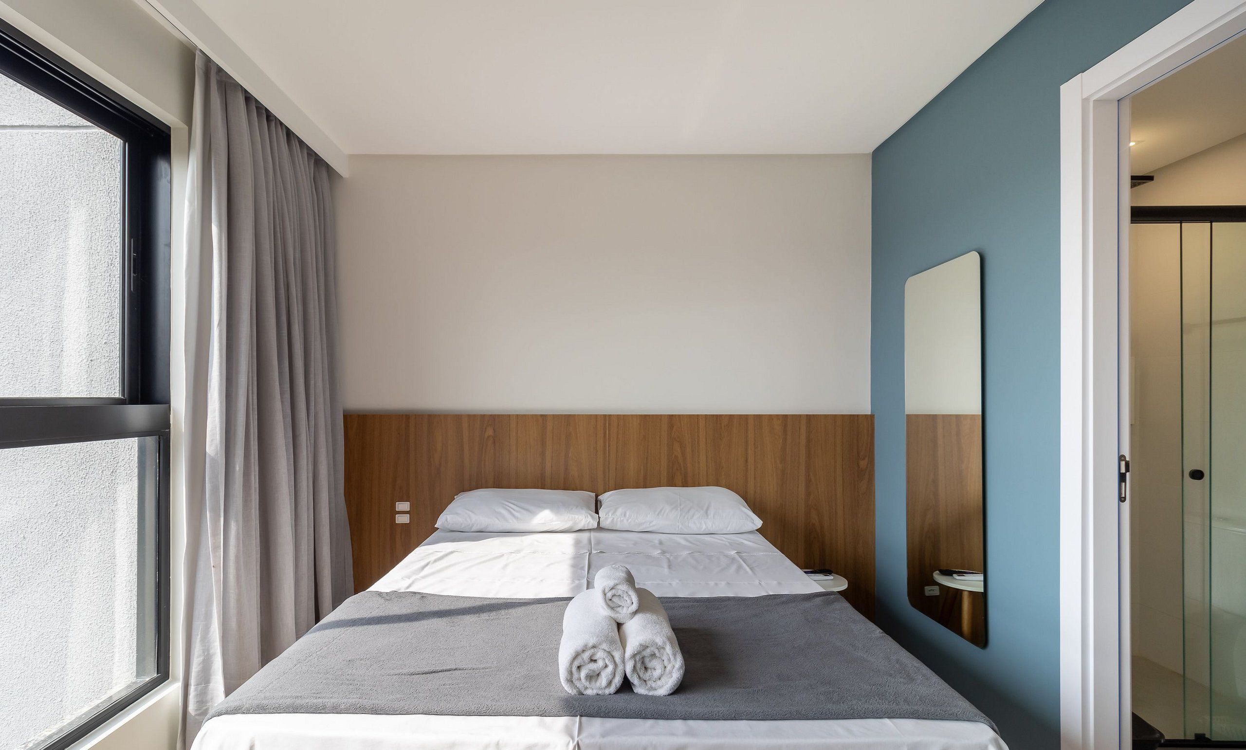 A hotel room with a bed, a window with curtains, a wooden headboard, a mirror, and a door.