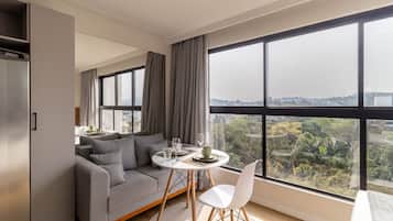 A modern living room with a grey sofa, a dining table with chairs, and a large window offering a city view.