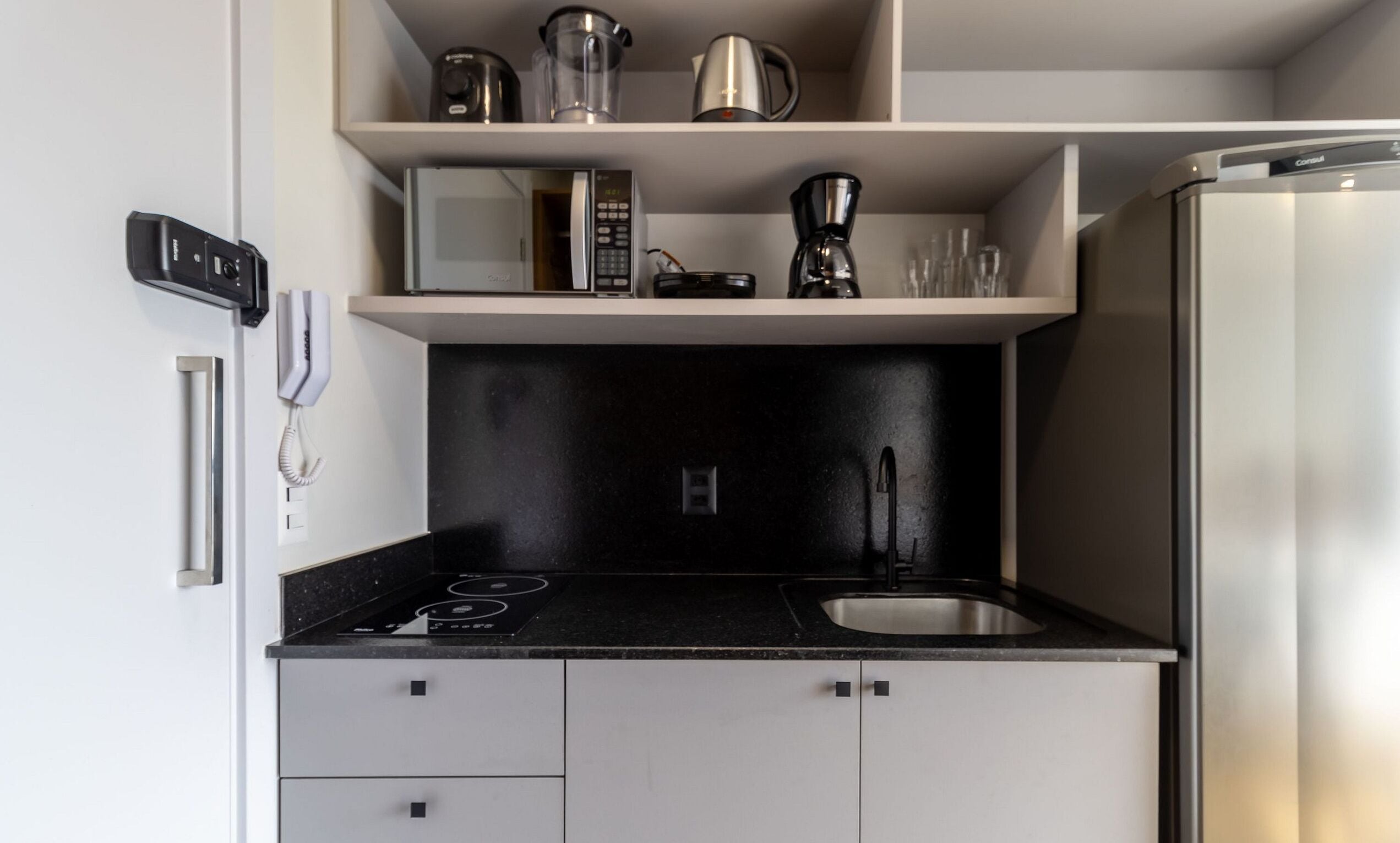 A compact kitchen with a black countertop, a stainless steel refrigerator, and a coffee maker on the shelf.
