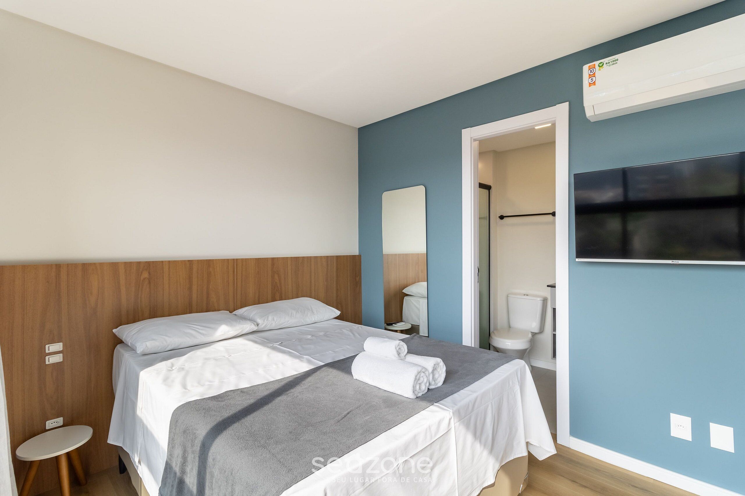 Quarto de hotel com cama, um banquinho, espelho, televisão e banheiro visível por uma porta aberta.