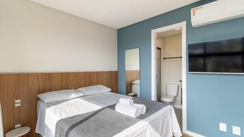 Quarto de hotel com cama, um banquinho, espelho, televisão e banheiro visível por uma porta aberta.