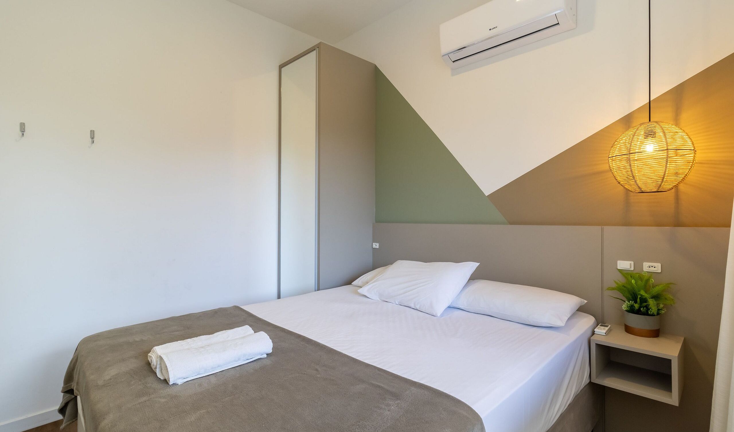 A hotel room with a bed, a bedside table with a plant, an air conditioning unit, and a wall-mounted light fixture.