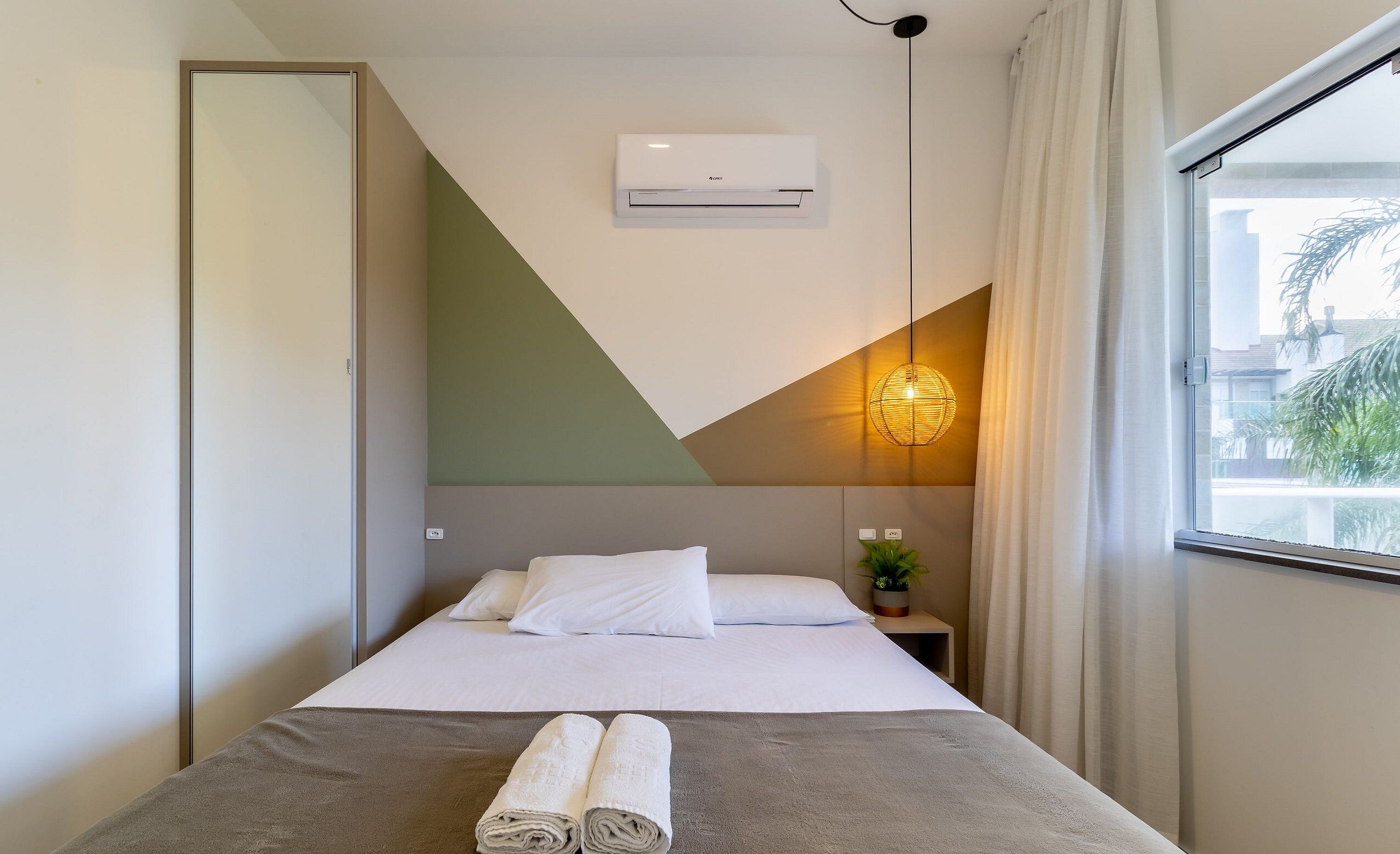 Ein Hotelzimmer mit Bett, Nachttisch mit Pflanze, Klimaanlage und einem Vorhangfenster.