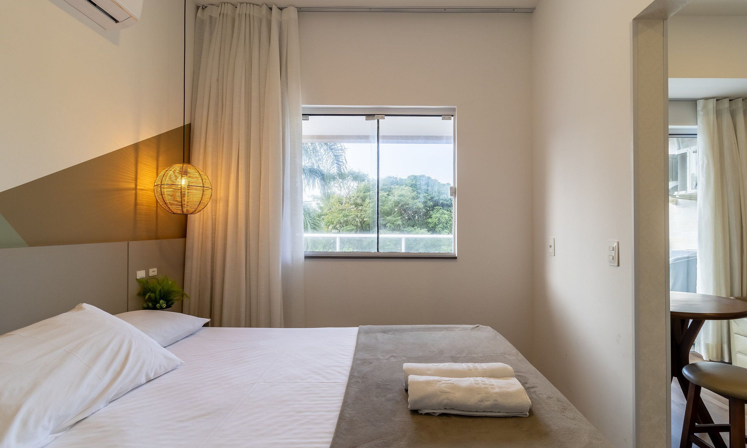 Ein Hotelzimmer mit Bett, Fenster mit Blick auf Grünflächen, Nachttisch mit Lampe und einem Sessel.