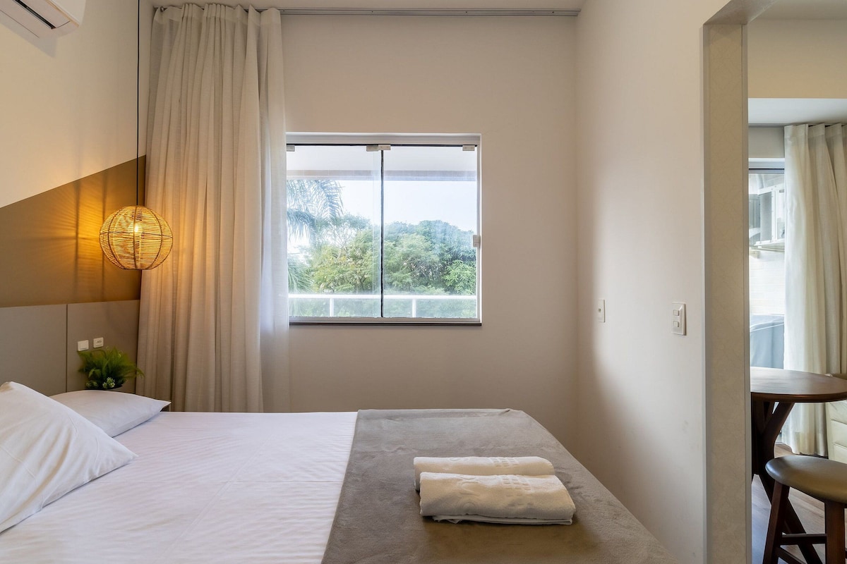 Ein Hotelzimmer mit Bett, Fenster mit Blick auf Grünflächen, Nachttisch mit Lampe und einem Sessel.