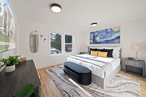 A modern bedroom with a large bed, a painting above the bed, a desk with a plant, and a window with a view.
